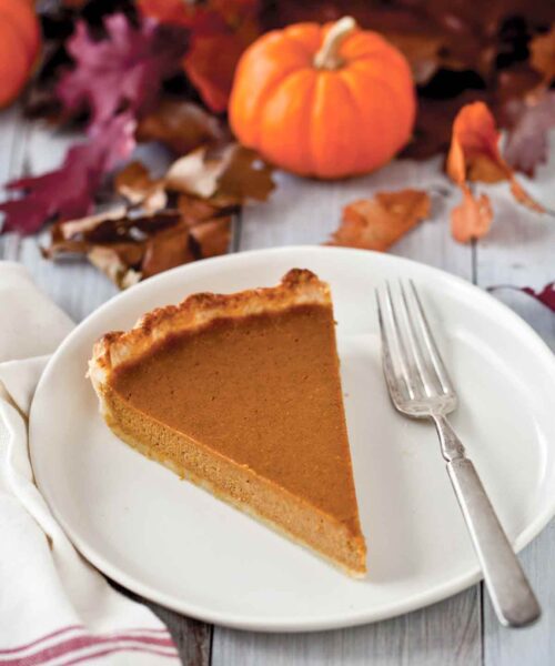 A slice of drunken pumpkin bourbon tart on a white plate with a fork beside it and a mini pumpkin and leaves in the background.