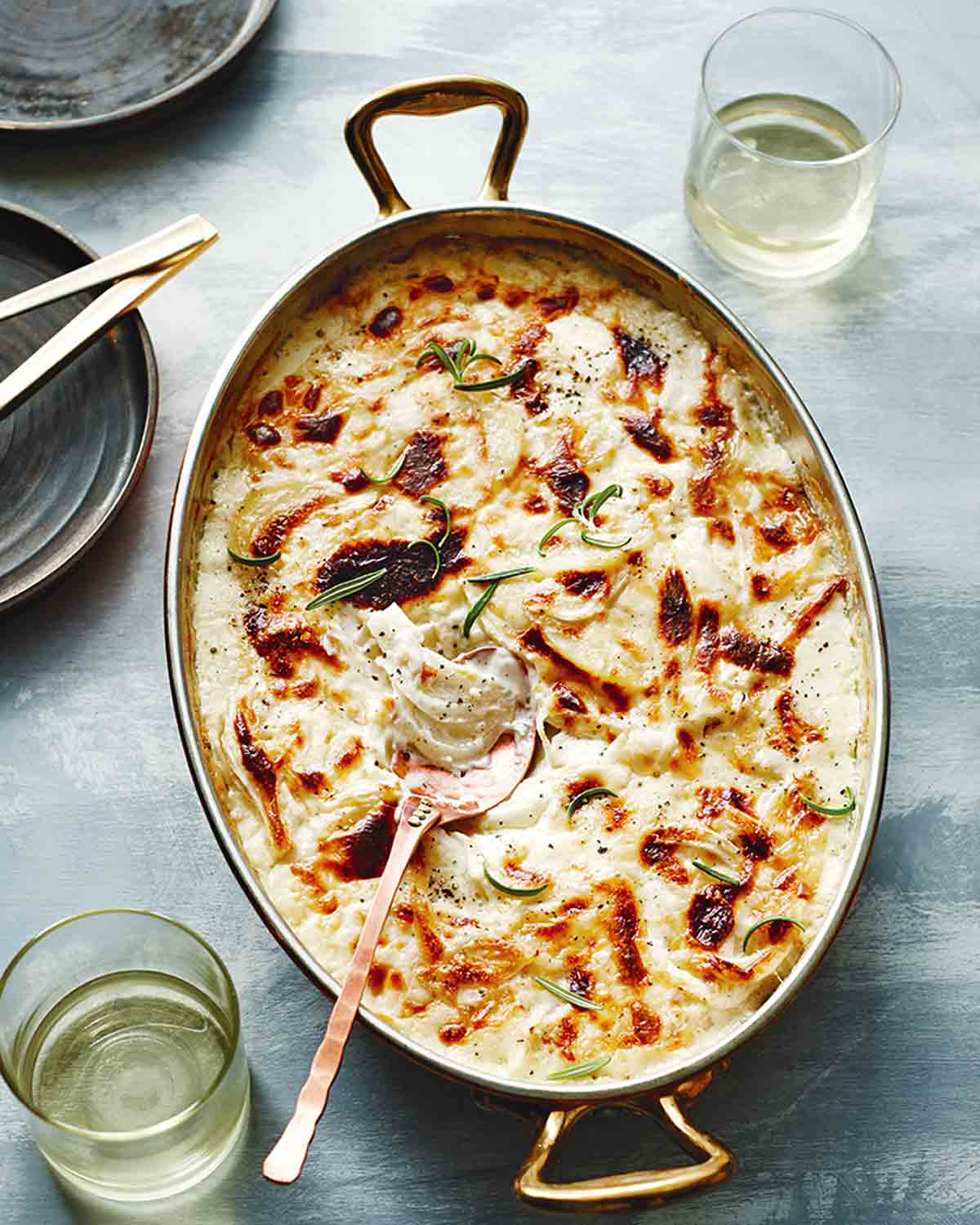 A browned fennel potato gratin, garnished with rosemary in an oval casserole dish with handles and a glass of white wine on either side of the dish.