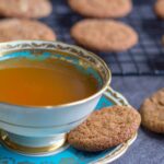 A molasses spice cookie on the saucer of a cup of tea, more cookies in a pile and on a wire rack.