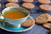 A molasses spice cookie on the saucer of a cup of tea, more cookies in a pile and on a wire rack.