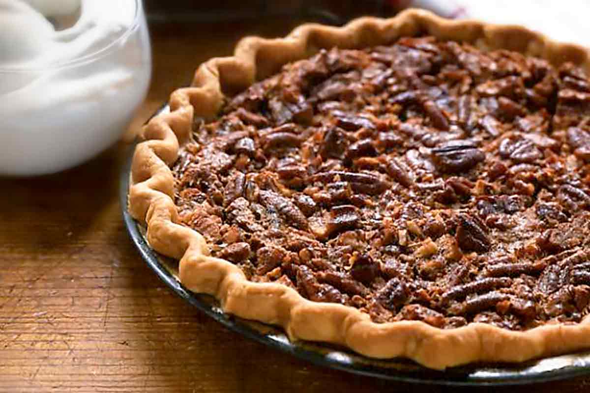 Two southern pecan pies in pie plates with a bowl of whipped cream between them.