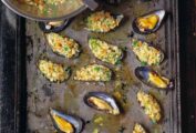 Several opened mussels on a baking sheet, some filled with stuffing and a bowl of extra stuffing on the side.
