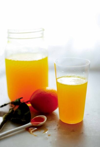 A mason jar and a tall glass filled with turmeric lemonade, and a spoon and orange lying nearby.