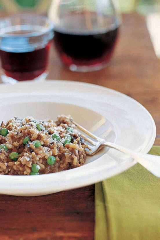 A white bowl of wild mushroom risotto with peas with a fork resting inside and a glass and decanter of wine in the background.