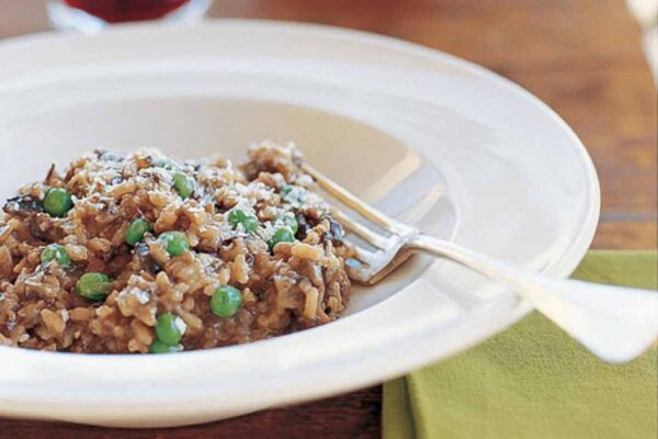 A white bowl of wild mushroom risotto with peas with a fork resting inside and a glass and decanter of wine in the background.