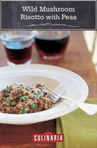 A white bowl of wild mushroom risotto with peas with a fork resting inside and a glass and decanter of wine in the background.