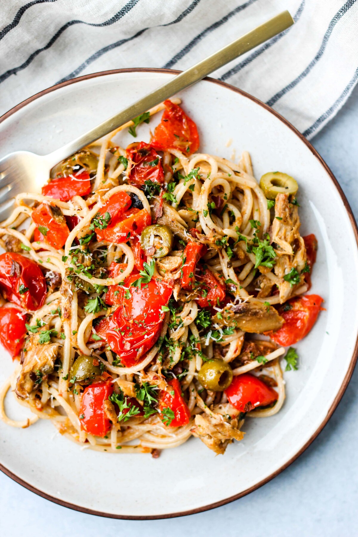 A white plate filled with pasta, canned mackerel, tomatoes, olives, and fresh parsley, with a gold fork on the side.