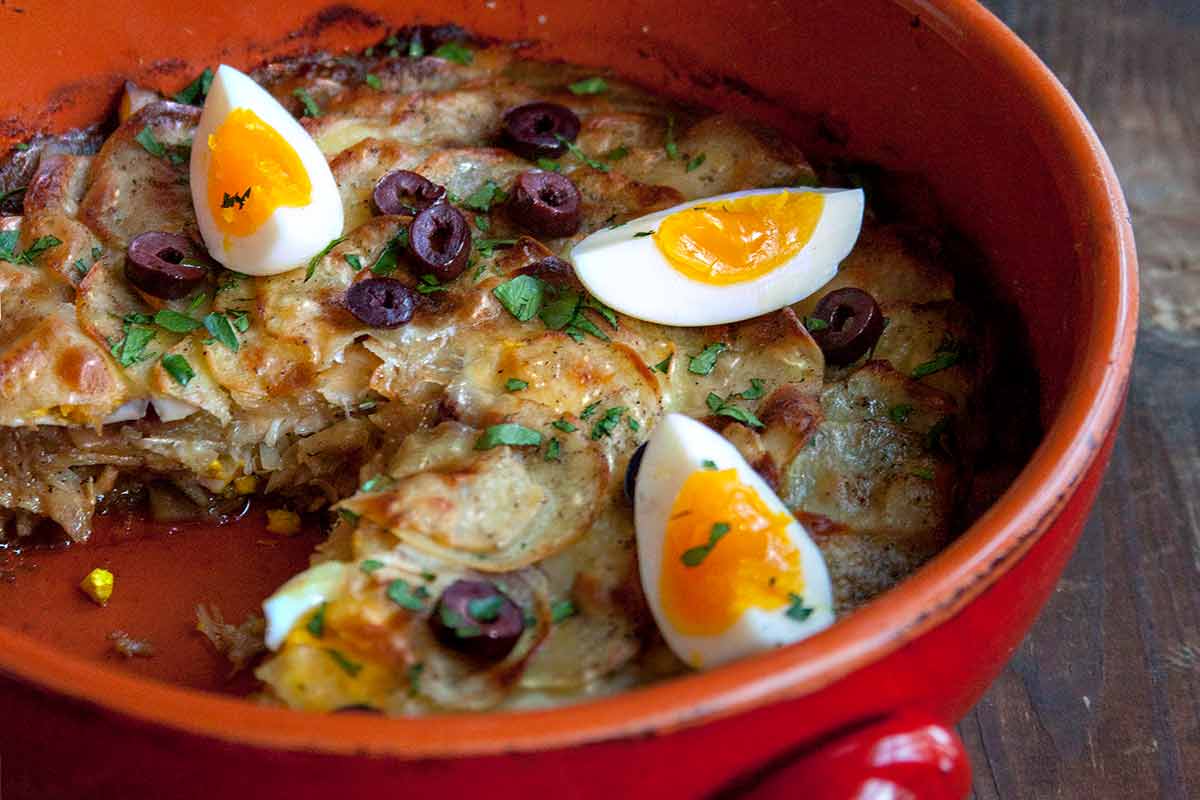 Crockery filled with bacalhau a Gomes de Sa--layers of salt cod, potato, and, onions; on top, sliced olives, quartered boiled eggs, parsley on wood.