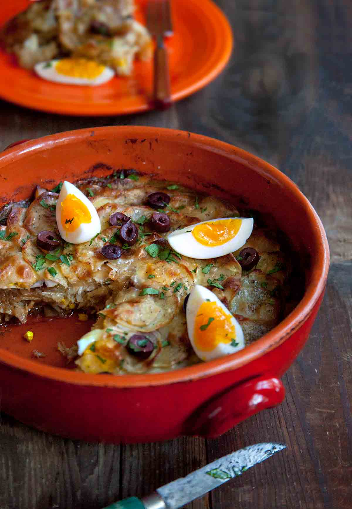 Crockery filled with bacalhau a Gomes de Sa--layers of salt cod, potato, and, onions; on top, sliced olives, quartered boiled eggs, parsley on wood.