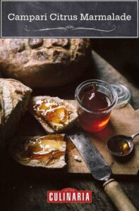 Two loaves of artisan bread and a jar of Campari citrus marmalade, with a slice of bread smeared with the marmalade on a wooden cutting board.