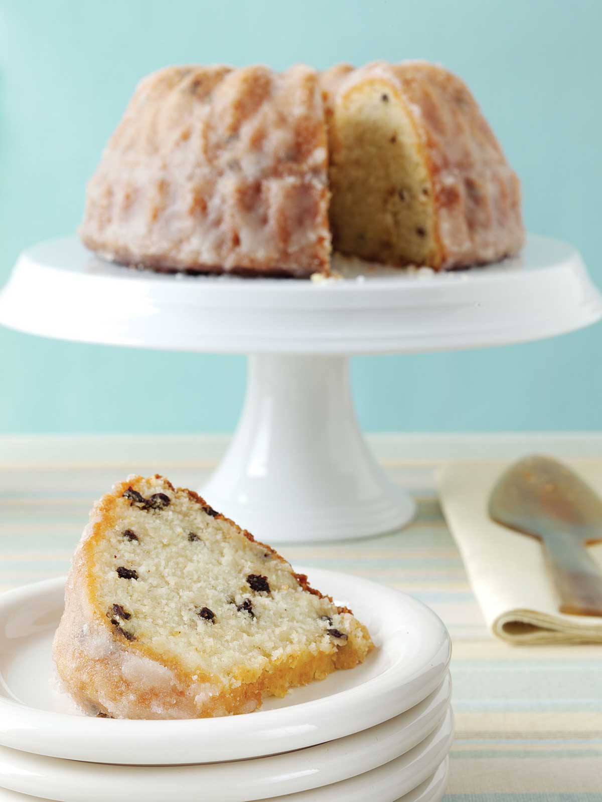 A plate with a slice of pound cake with currants, and a cake stand in the background with the remaining cake.