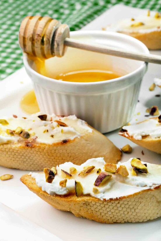 Toasted rounds of baguette topped with goat cheese and chopped pistachios with a dish of honey in the background.