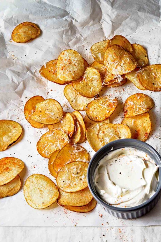 Homemade potato chips on a crumpled piece of parchment paper and a bowl of sour cream.