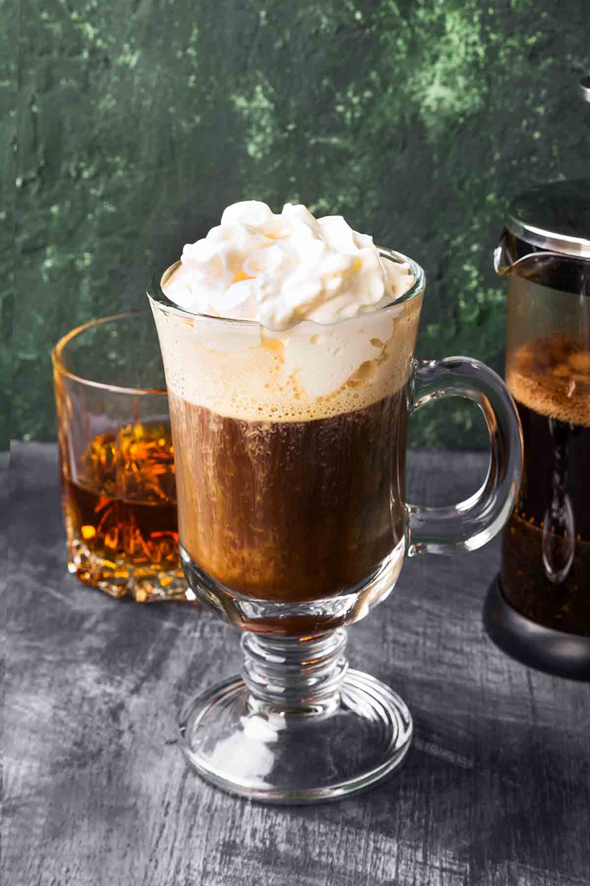 A tall glass mug filled with Irish coffee topped with whipped cream and a tumbler or whiskey in the background.