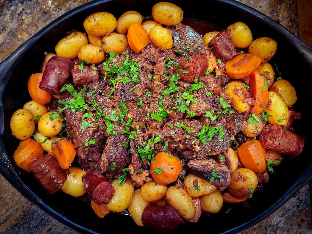 A black platter filled with slow cooker carne assada--beef chunks, carrots, potatoes, and sausage in a red broth.