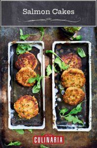 Six salmon cakes on rectangular platters with bowls of avocado spread and chimichurri beside.