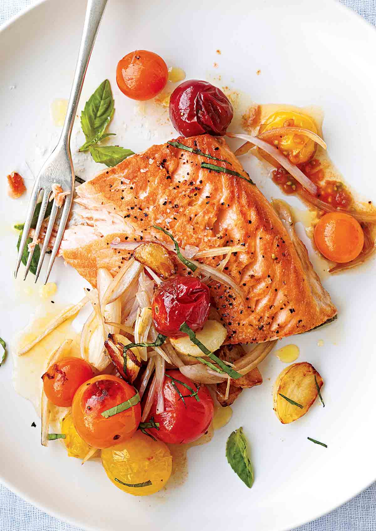 A white plate topped with a piece of cooked salmon with blistered cherry tomatoes, shallots, and fresh basil.