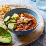 En skål med fläsk, avokado och hominy i en röd buljong på en skärbräda med tortillachips.