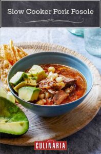 A bowl of pork, avocado, and hominy in a red broth on a cutting board with tortilla chips.