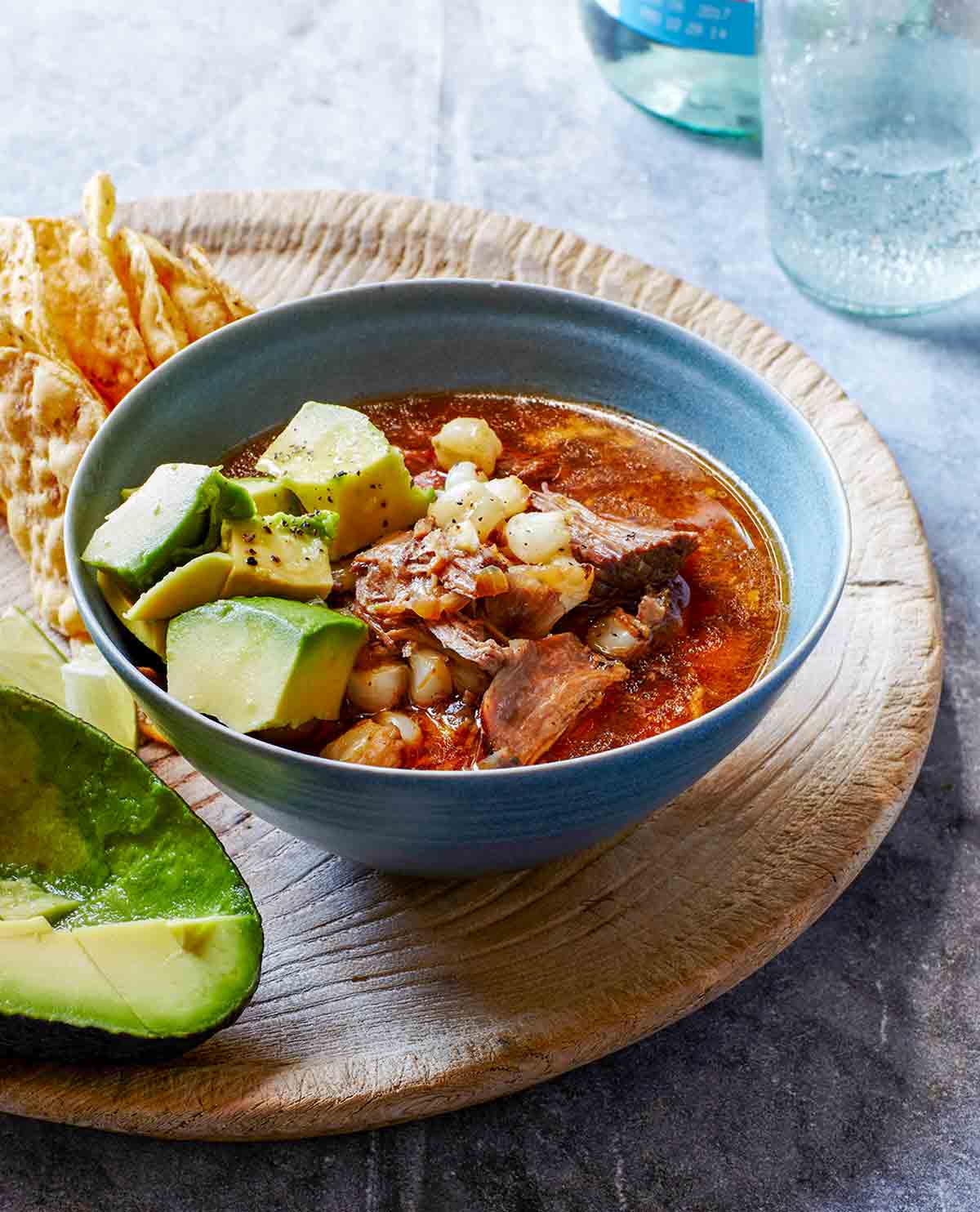 A bowl of pork, avocado, and hominy in a red broth on a cutting board with tortilla chips.