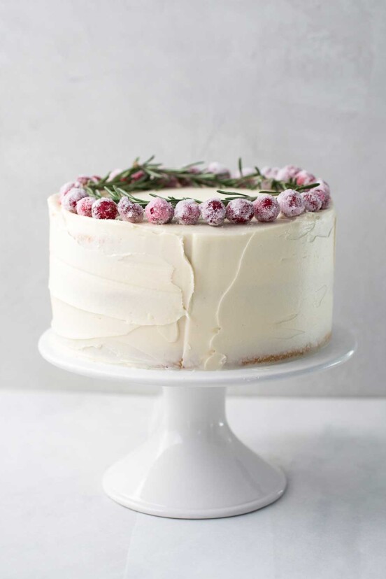 A whole frosted white Christmas cake topped with sugared cranberries and rosemary sprigs on a white cake stand.
