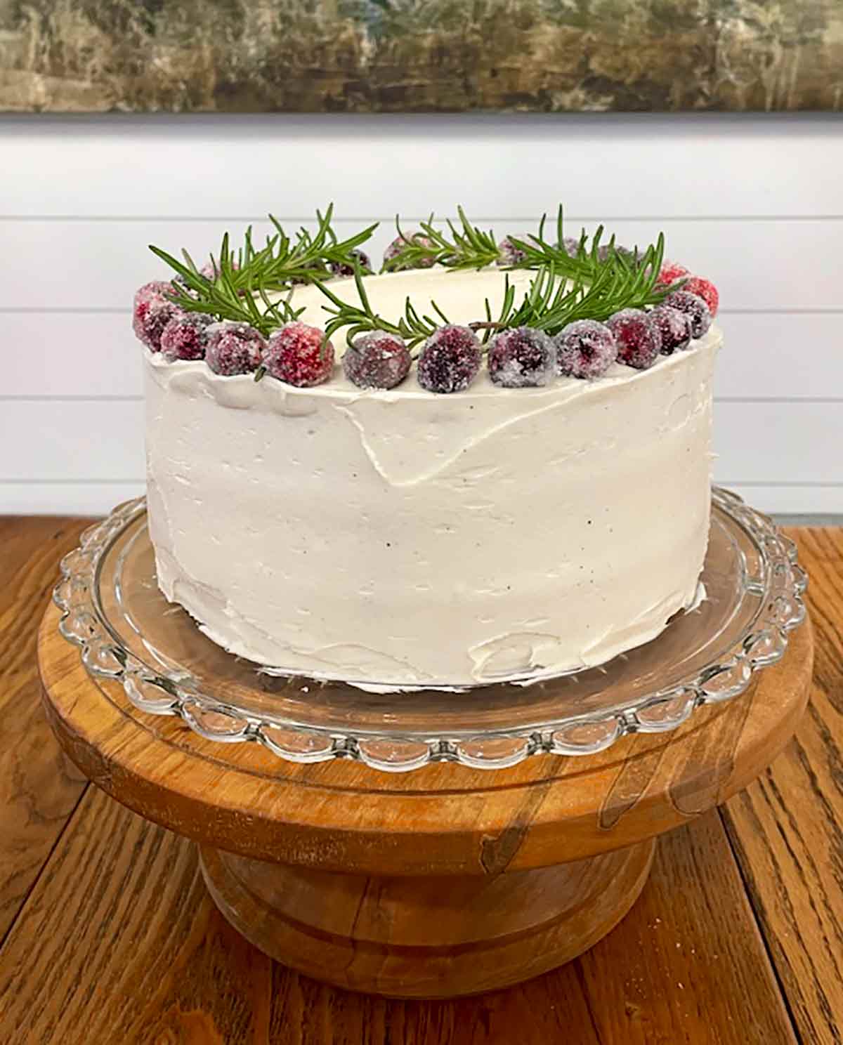 A whole frosted white Christmas cake topped with sugared cranberries and rosemary sprigs on a glass plate on a wooden cake stand.