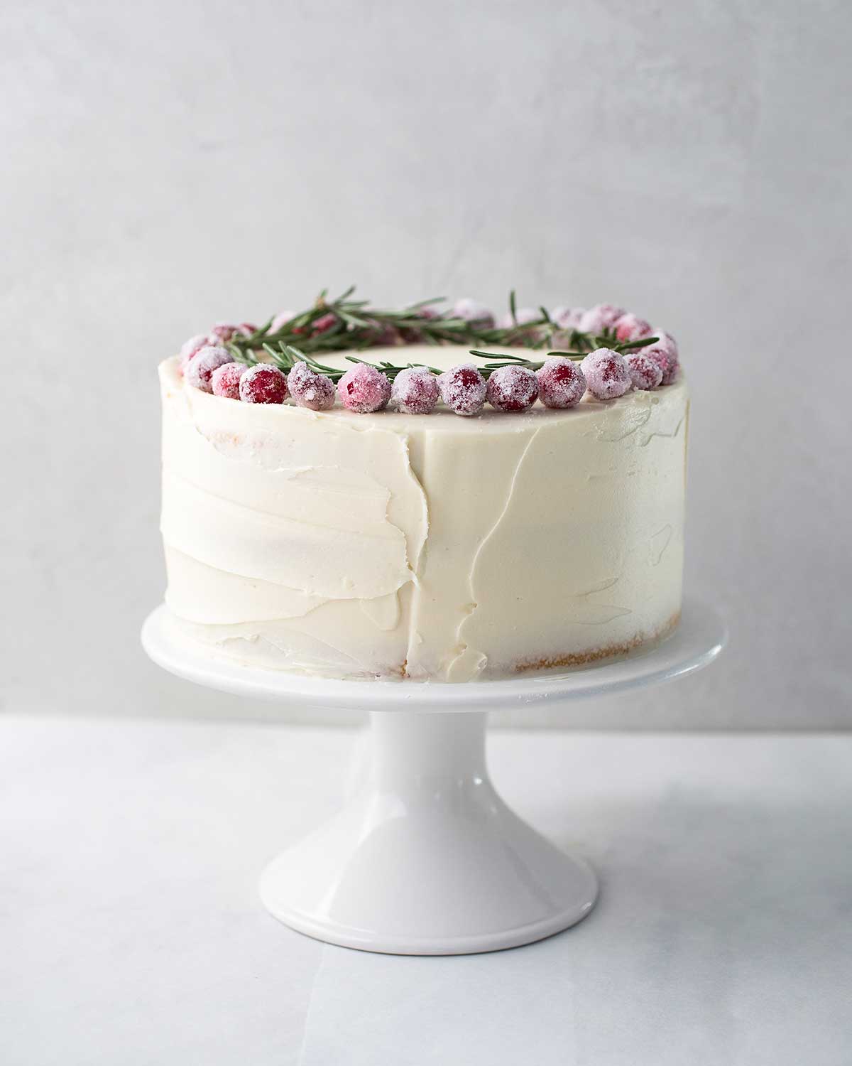 A whole frosted white Christmas cake topped with sugared cranberries and rosemary sprigs on a white cake stand.