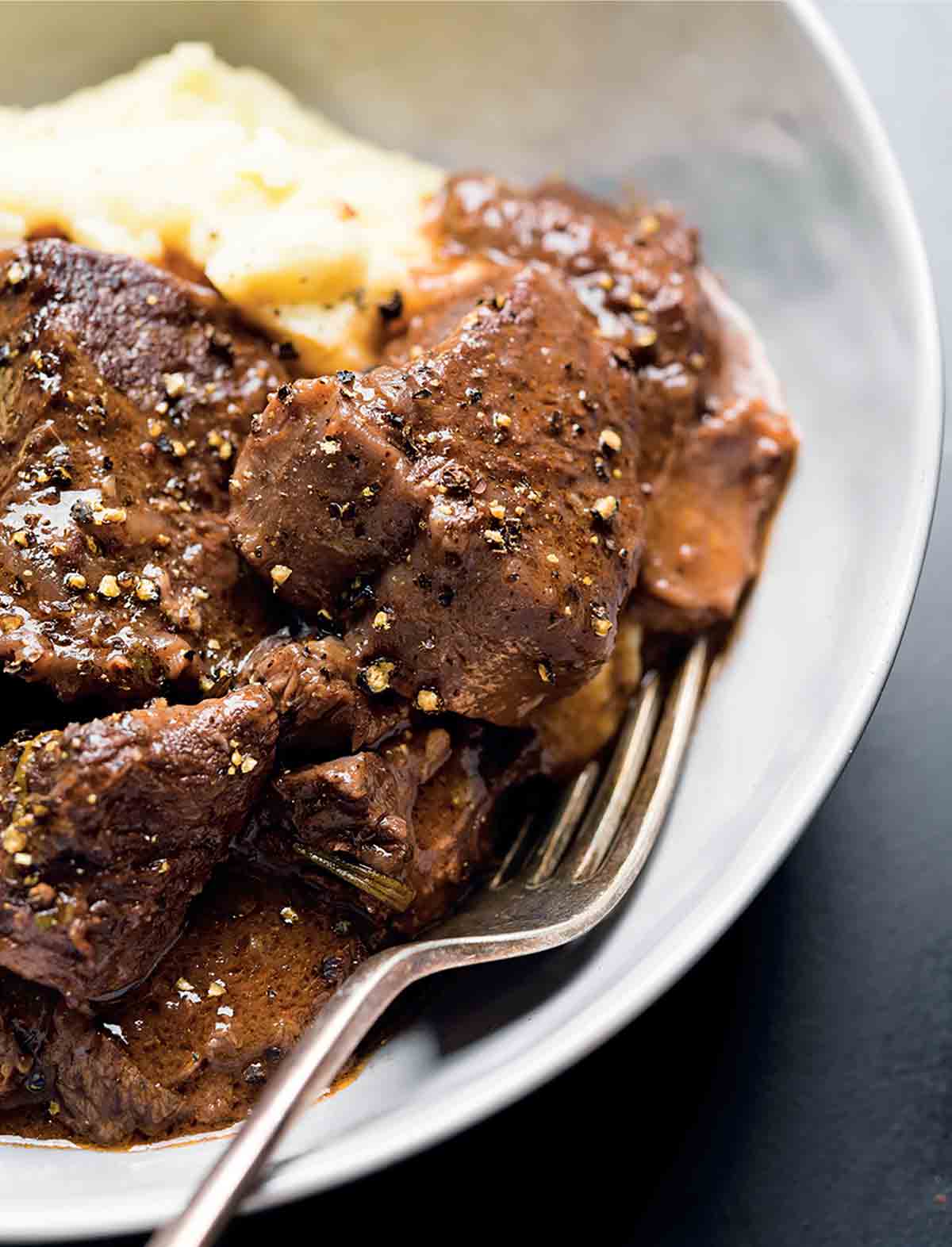 Pieces of beef stew with red wine and mashed potatoes in a white bowl with a fork nestled inside.