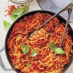 En stekpanna fylld med spagetti och köttsås med en tång och två basilikakvistar ovanpå.