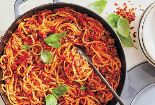 A skillet filled with spaghetti and meat sauce with a pair of tongs and two basil sprigs on top.