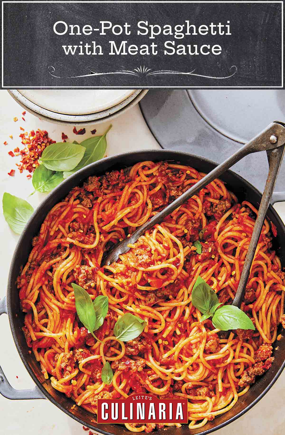 A skillet filled with spaghetti and meat sauce with a pair of tongs and two basil sprigs on top.