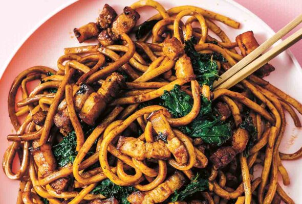 A pink bowl filled with fried Shanghai noodles, crispy pork belly, and kale, with chopsticks resting on the side.