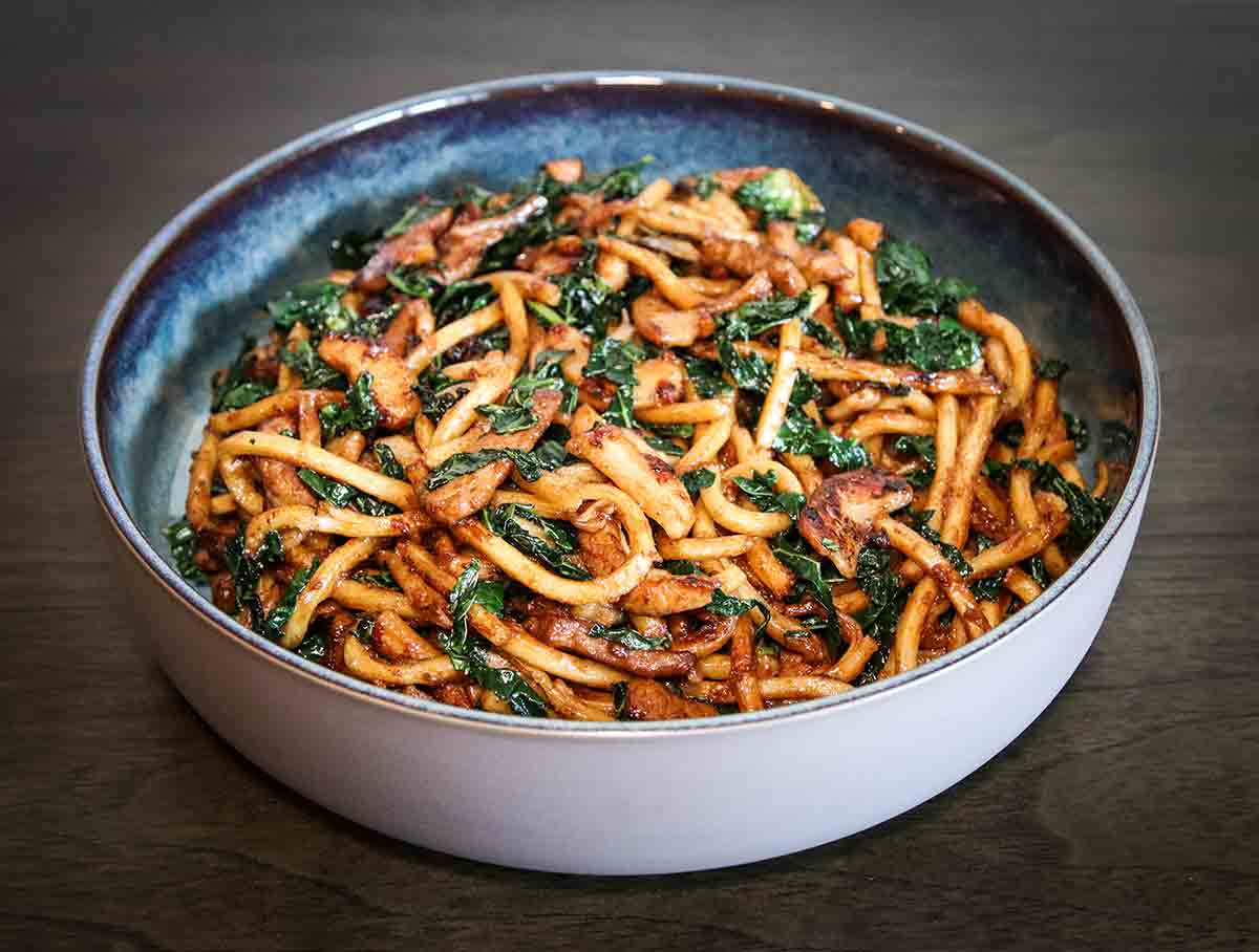 A white bowl filled with fried Shanghai noodles, crispy pork belly, and kale.