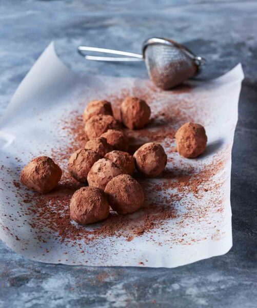 A sheet of parchment paper topped with chocolate truffles and a sifter of cocoa powder on the side.