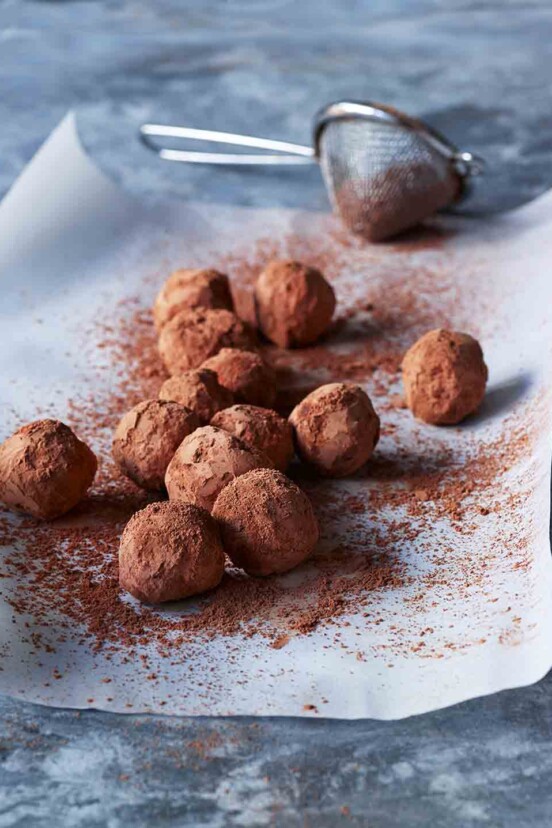 A sheet of parchment paper topped with chocolate truffles and a sifter of cocoa powder on the side.