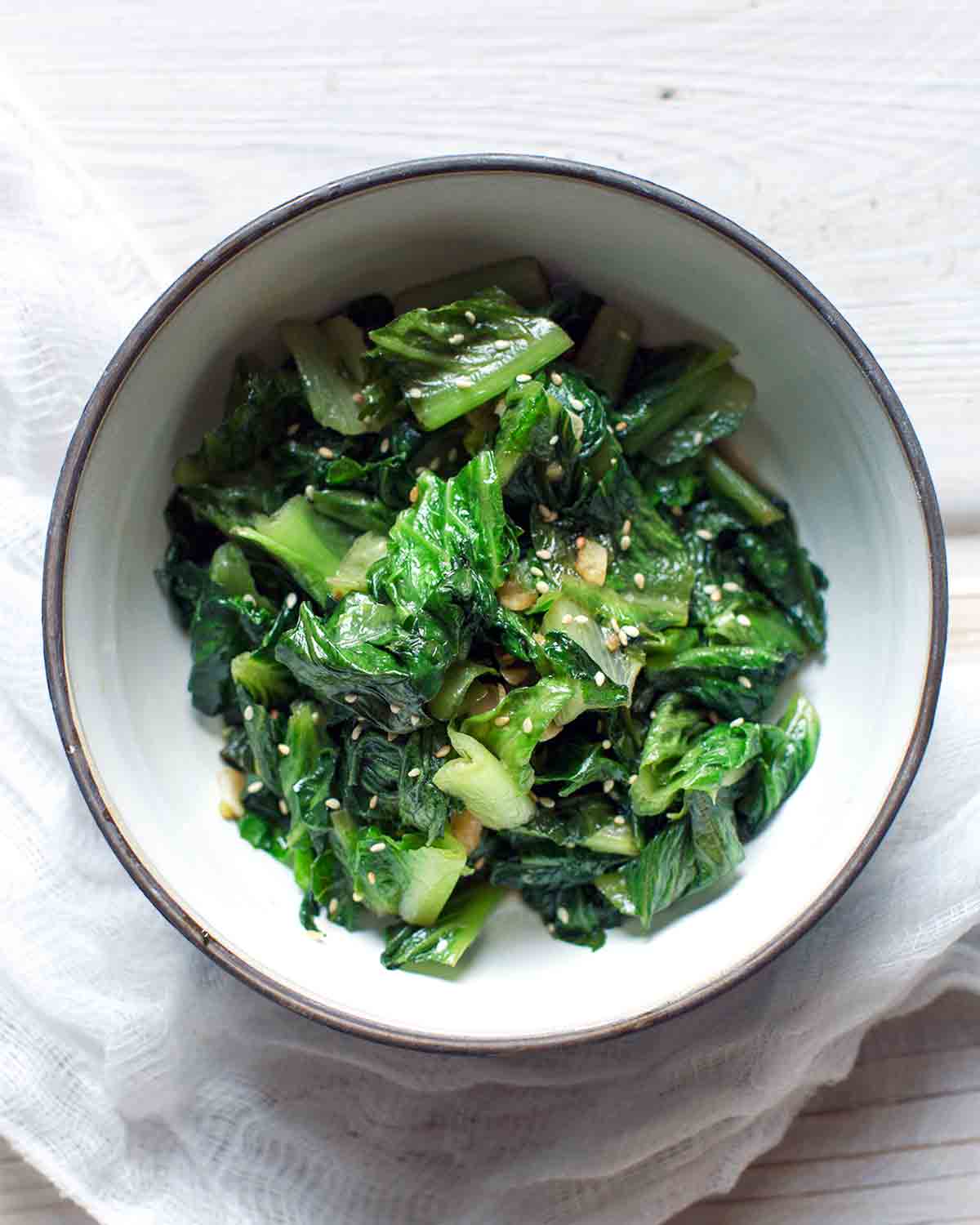 A white bowl filled with stir-fried lettuce, topped with sesame seeds on a white wooden surface.