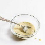 A glass bowl partially filled with creamy Italian salad dressing with a spoon resting inside.
