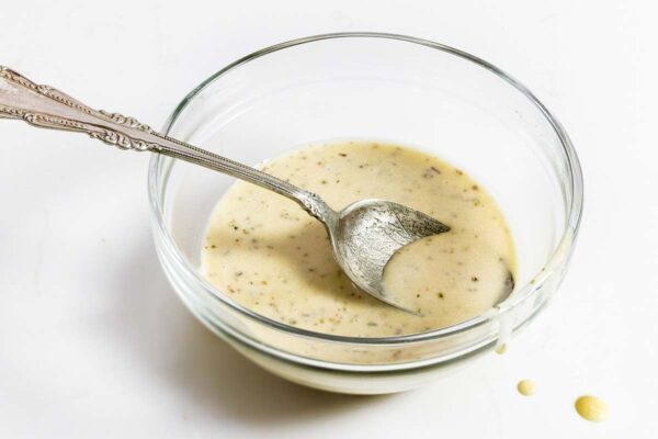 A glass bowl partially filled with creamy Italian salad dressing with a spoon resting inside.