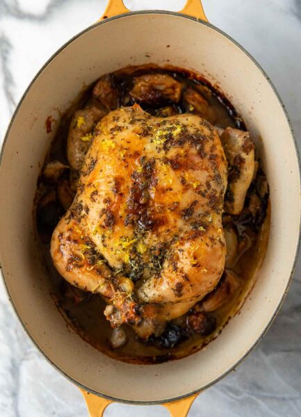 A large yellow Dutch oven with a roast chicken on a bed of cooked shallots, topped with lemon zest and herbs.