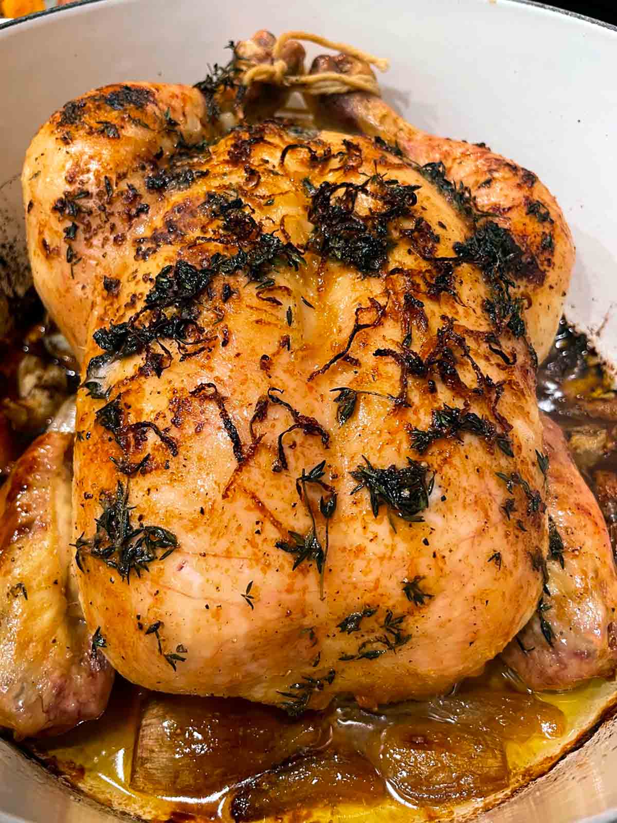 A large Dutch oven with a roast chicken on a bed of cooked shallots, topped with lemon zest and herbs.