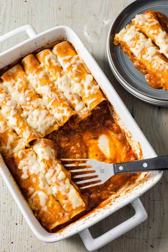 A rectangular dish of easy chicken enchiladas with a spatula resting in the dish and a couple enchiladas on a grey plate beside it.