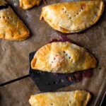 Five hand pies on a parchment-lined baking sheet with a spatula lifting one off.