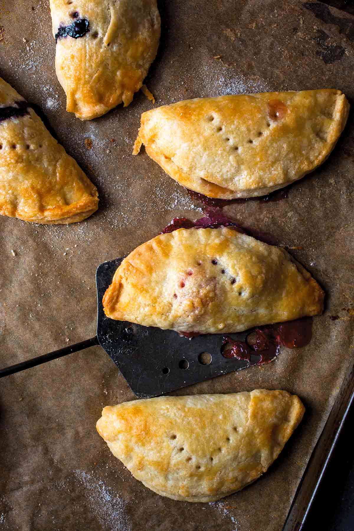 Five hand pies on a parchment-lined baking sheet with a spatula lifting one off.