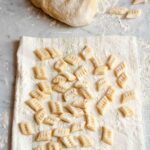Many prepared potato gnocchi on a floured kitchen towel, with a mound of gnocchi dough in the background.