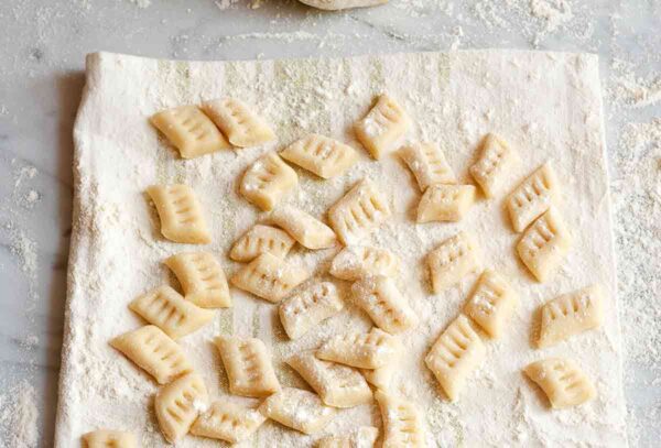 Many prepared potato gnocchi on a floured kitchen towel, with a mound of gnocchi dough in the background.