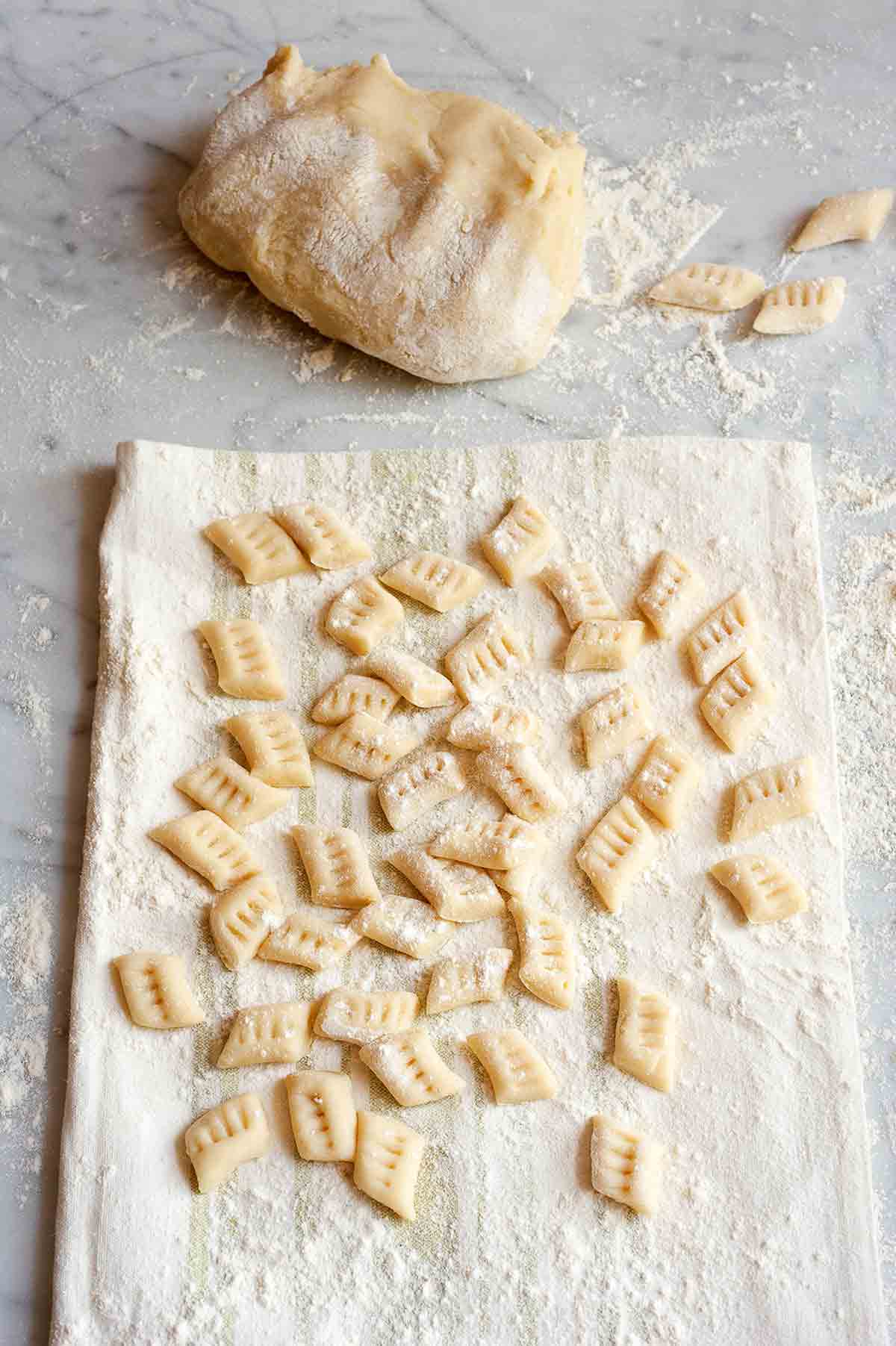 Many prepared potato gnocchi on a floured kitchen towel, with a mound of gnocchi dough in the background.