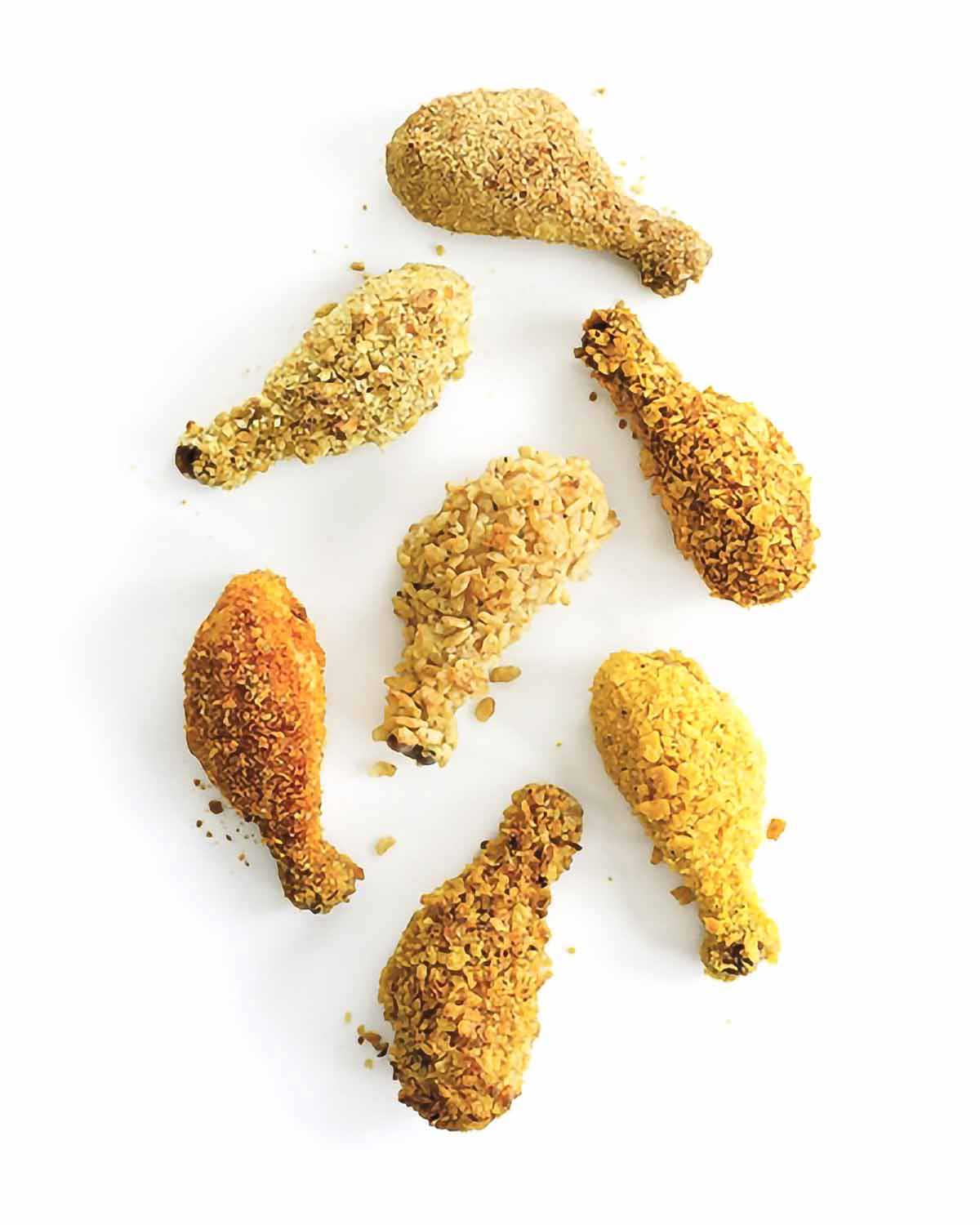 Seven oven fried drumsticks with different coatings on a white background.