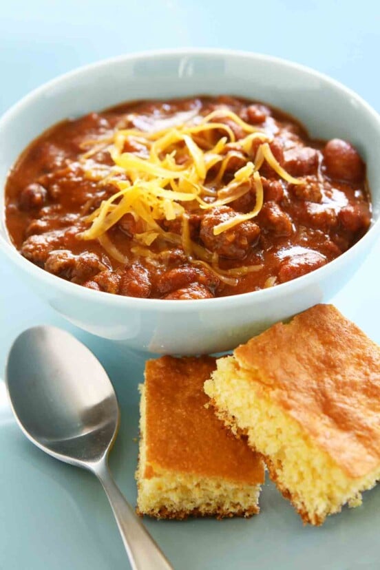 A white bowl filled with chili, with shredded cheese on top, and a spoon and two pieces of cornbread on the side.