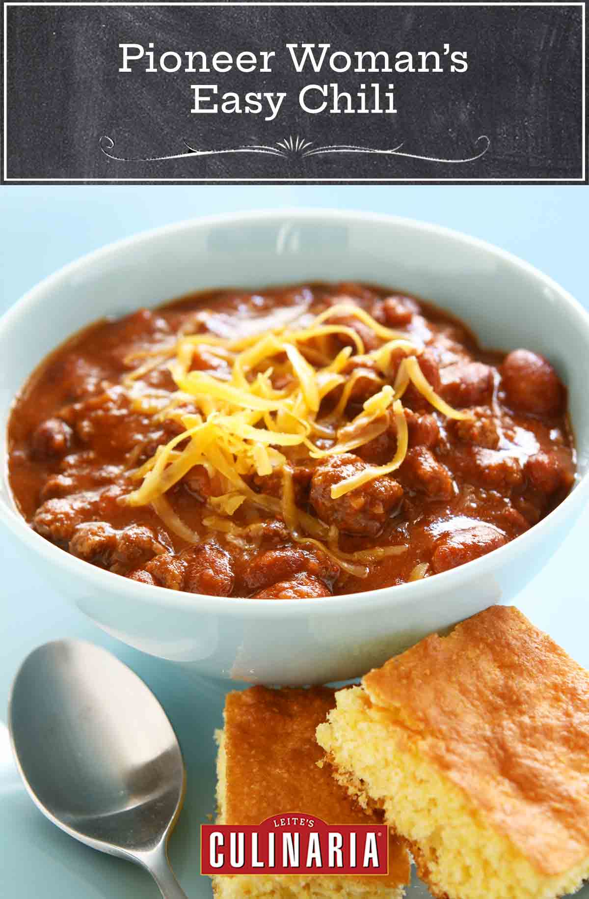 A white bowl filled with chili, with shredded cheese on top, and a spoon and two pieces of cornbread on the side.