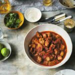 A bowl of pork tinga--pork stewed with tomatoes and chipotle peppers, along with bowls of lemon, sour cream, cilantro, cheese on a table.